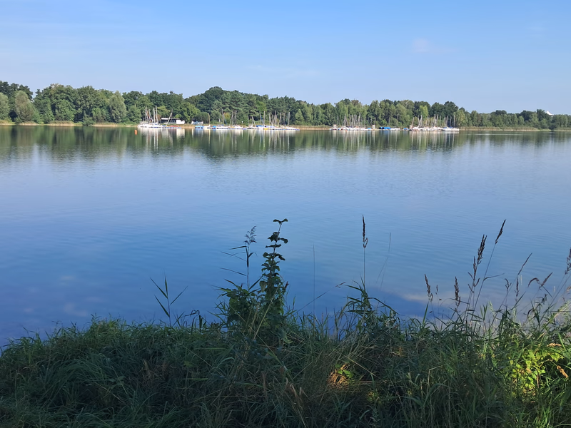 Ein ruhiger See und blauer Himmel, im Hintergrund Bootsstege und Boote