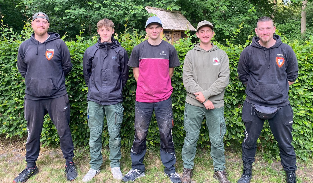 Drei Jugendliche, flankiert von zwei Erwachsenen, stehen vor einer grünen Hecke.