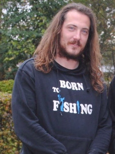 <p>Ein Mann mit langen Haaren trägt schwarzen Kapuzenpullover mit weißem Text 'BORN TO FISHING' und blauen Fischmotiven. Er steht vor einer Hecke und Büschen.</p>
