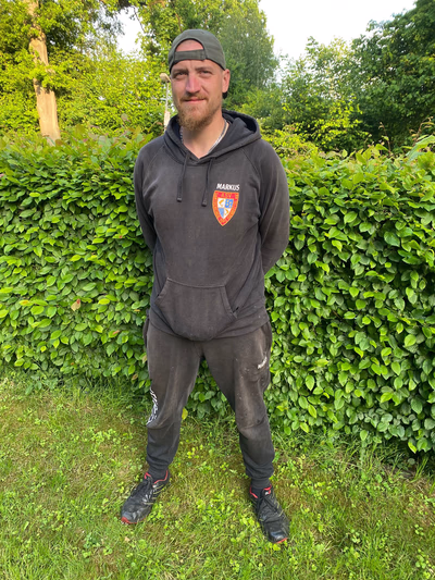 Mann in schwarzem Kapuzenpullover mit Wappen und schwarzer Anglerjogginghose mit Stiefeln, stehend vor einer grünen Hecke. Er trägt ein Kappi, was er verkehrt herum aufgesetzt hat.