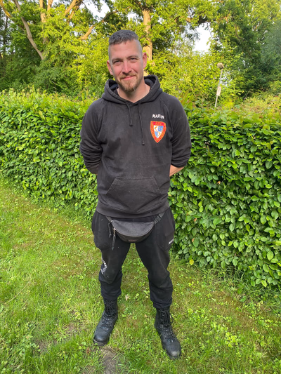 Mann in schwarzem Kapuzenpullover mit Wappen und schwarzer Hose mit Stiefeln, stehend vor einer grünen Heckereins.